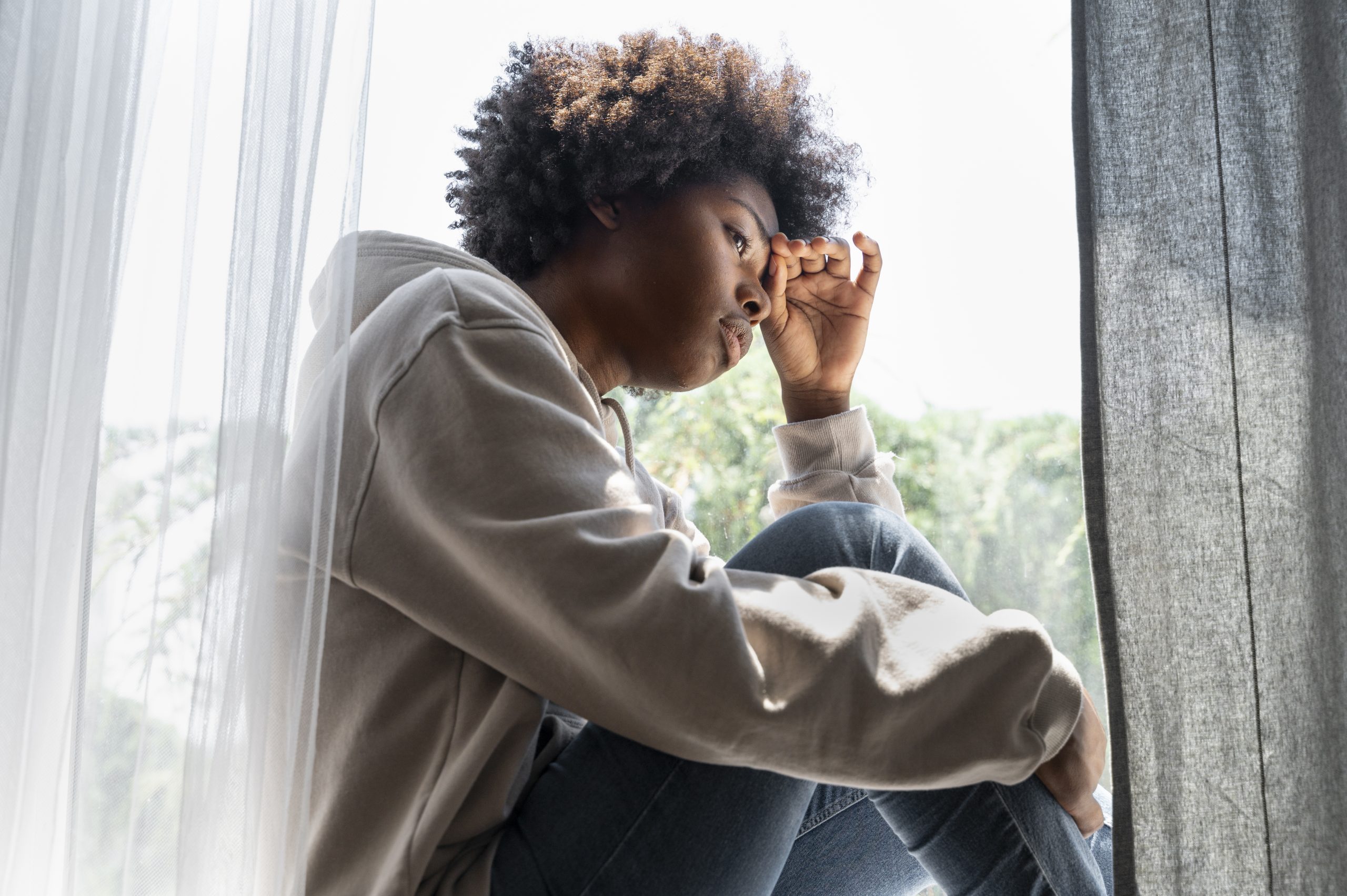 A person in a hoodie sitting by a window with one hand over their eyes.
